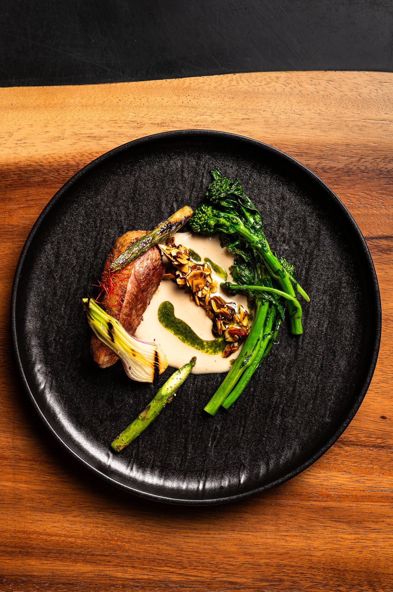 duck breast, asparagus, grilled leeks, broccolini, almonds, creamy sauce with herb oil served on a black fine-dining plate on a wooden table