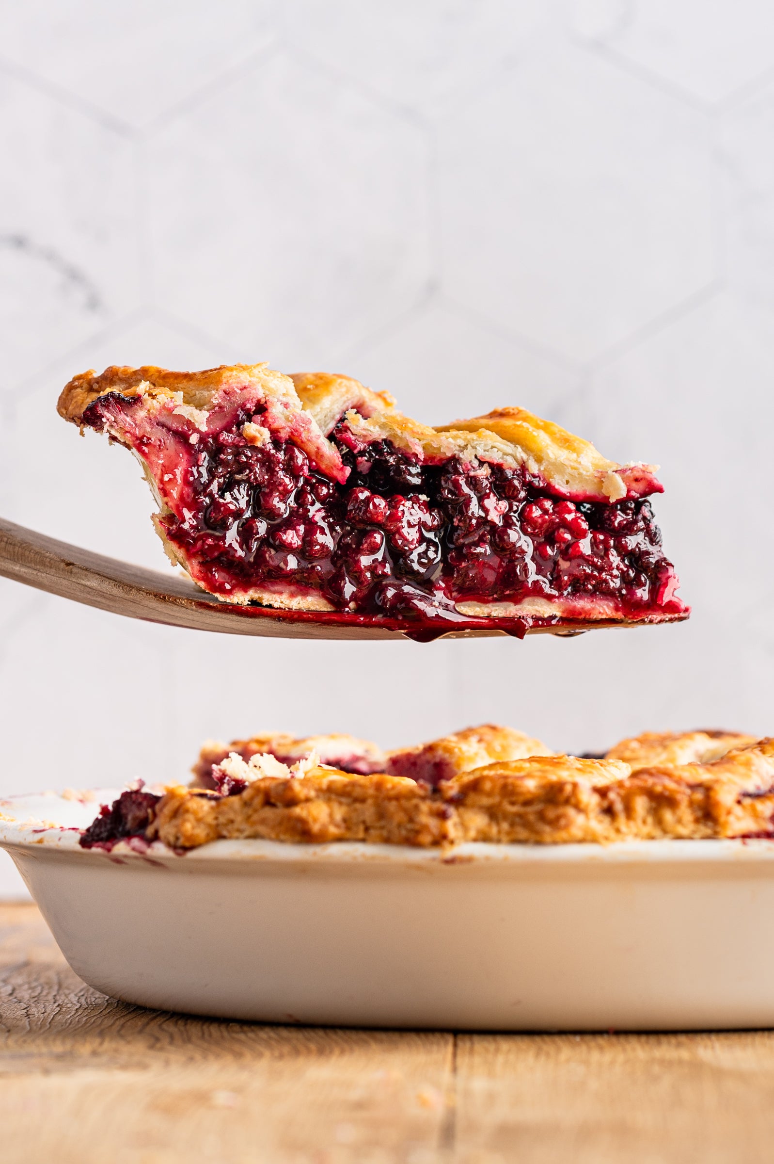 Slice of berry pie being lifted from a whole pie on a wooden board.