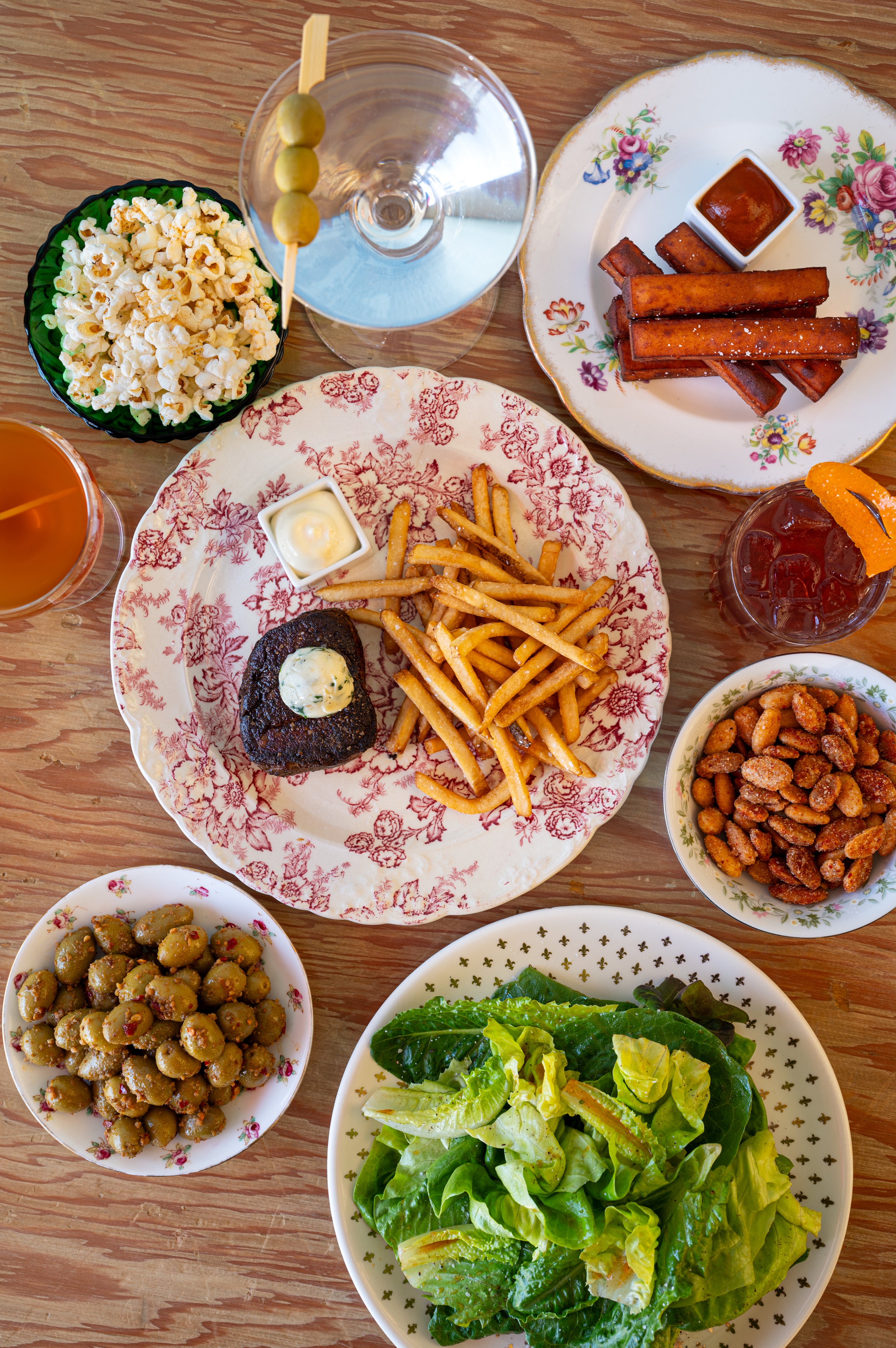 flatlay of contemporary French food featuring steak frites, spiced nuts, salad, olives, popcorn, polenta fries, and alcoholic drinks