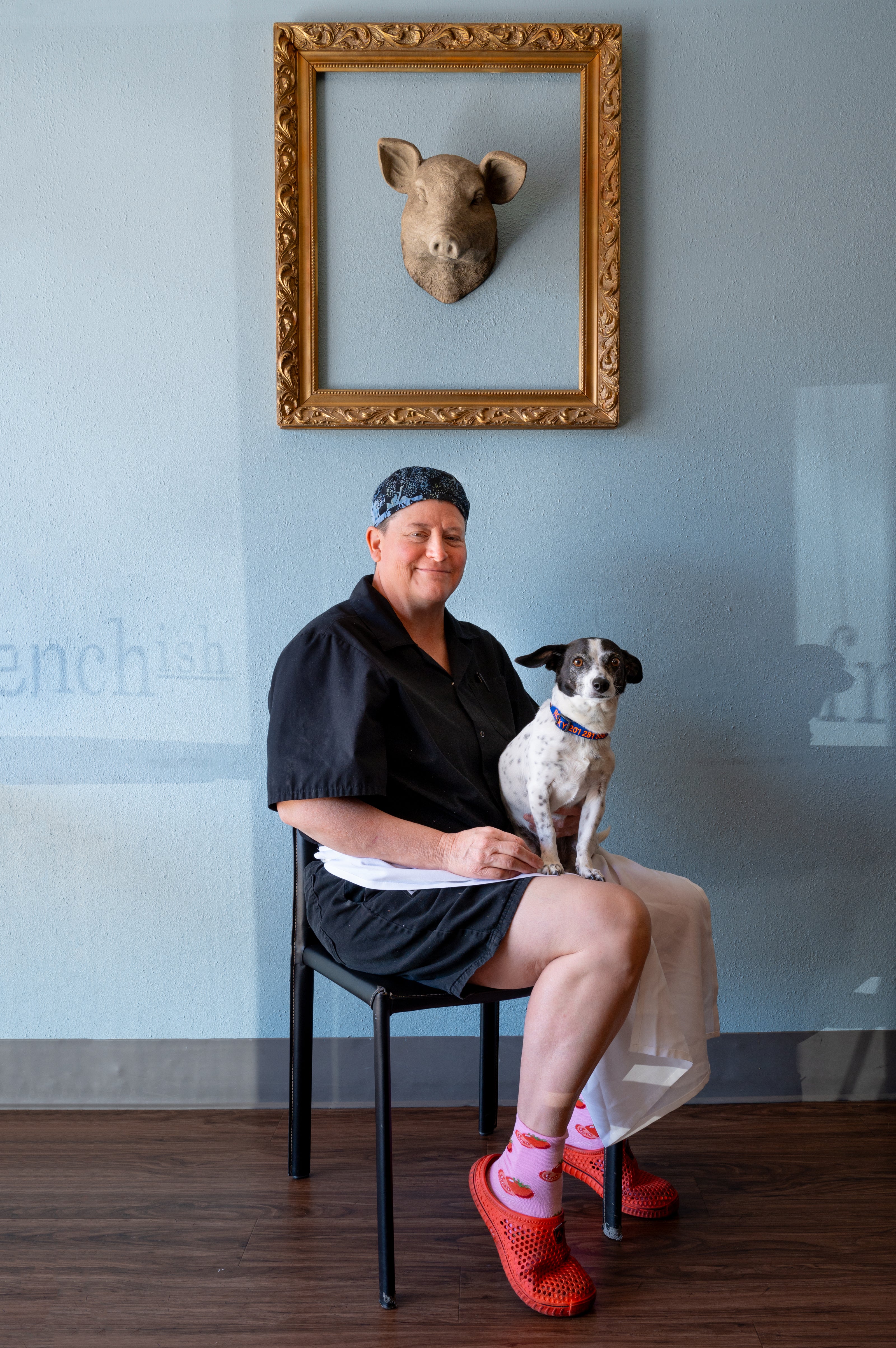 restaurant owner in chef clothes sitting down holding their dog underneath a fake pigs head with a frame surrounding it