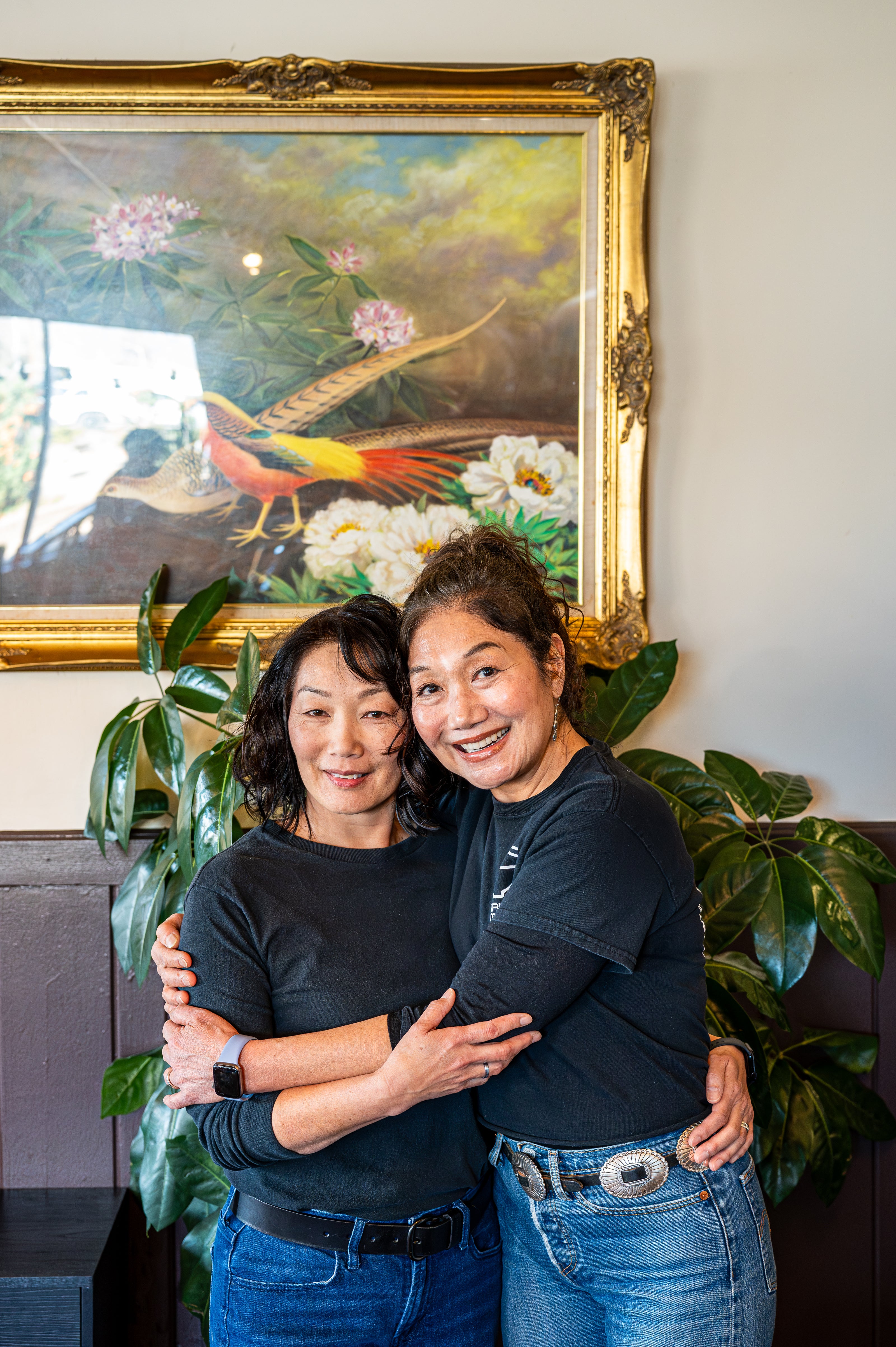 portrait of ramen restaurant owners inside their restaurant hugging each other under a painting from their relative