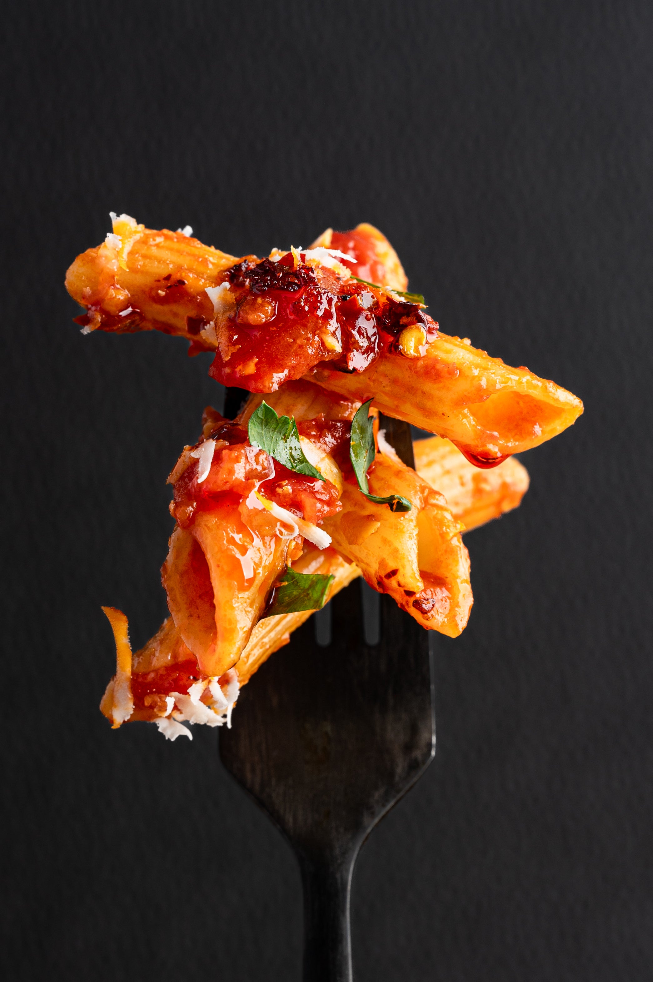 fork holding pieces of penne arrabiata using chili crisp and garnished with grated parmesan cheese and parsley with a dark background