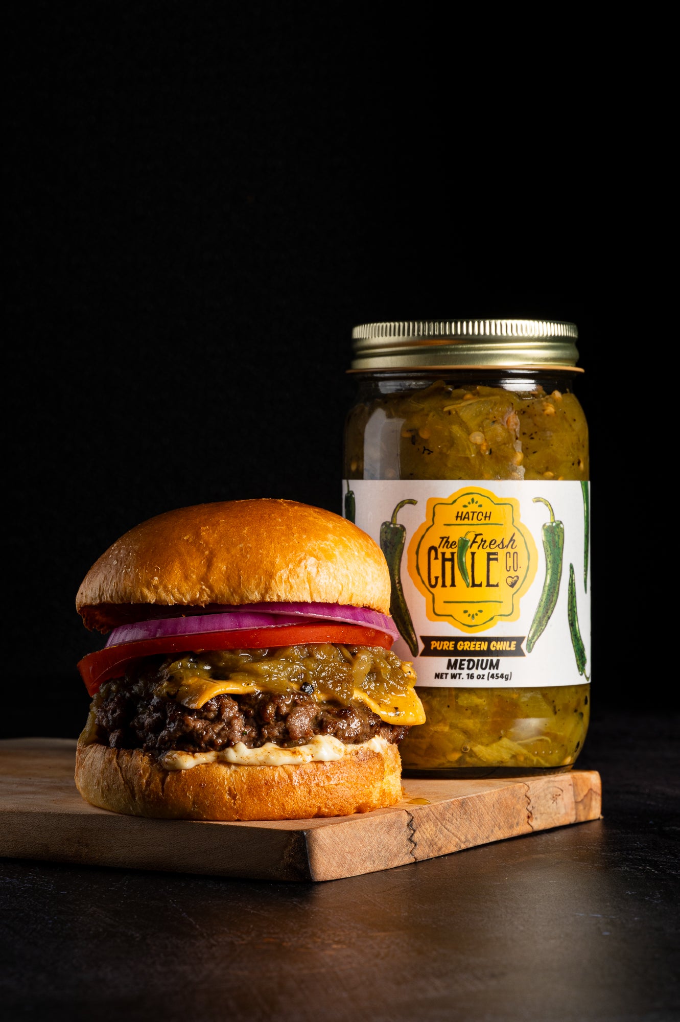 green chile cheeseburger on a wooden cutting board with a jar of pure Hatch green chile with a dark background