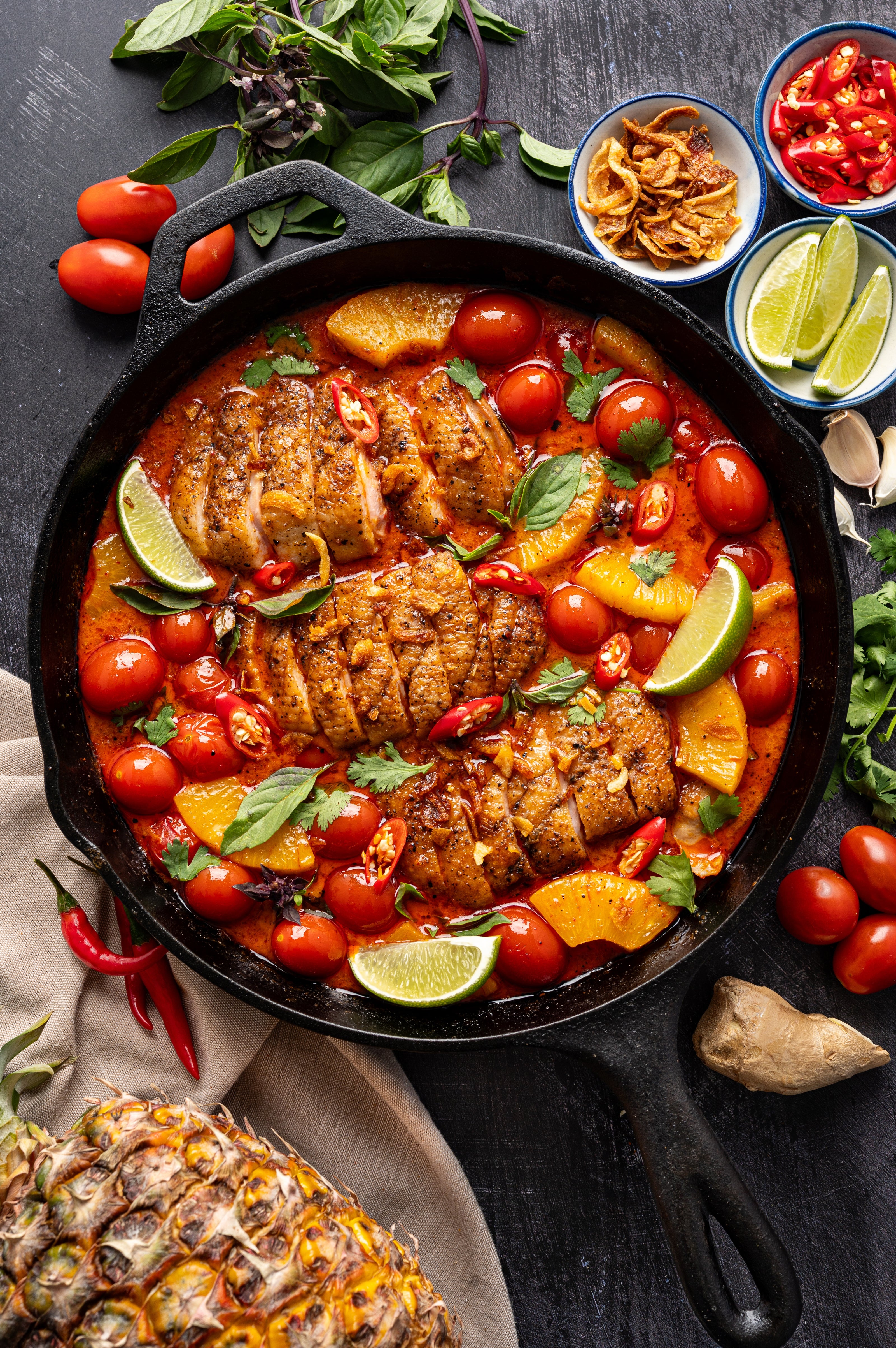 Thai red curry in a cast iron pan with cherry tomatoes, pineapple, Thai chiles, cilantro, ginger, lime, crispy shallots on a dark background